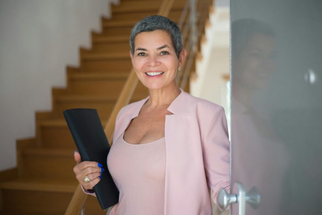 woman with salt and pepper pixie cut wearing pink shirt and open pink blazer, smiling at cameray olding a black portfolio with stairs behind her and leaning on a door