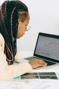 side view of woman with black pink and green braids and wearing glasses and a cream colored suit. sitting at a table looking at graphs on a laptop